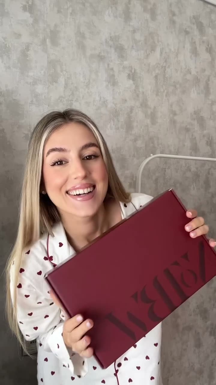 A vertical direct-to-camera unboxing: a creator holds up the brand's maroon wordmark box with a big smile, then tries on the pink-and-green set.